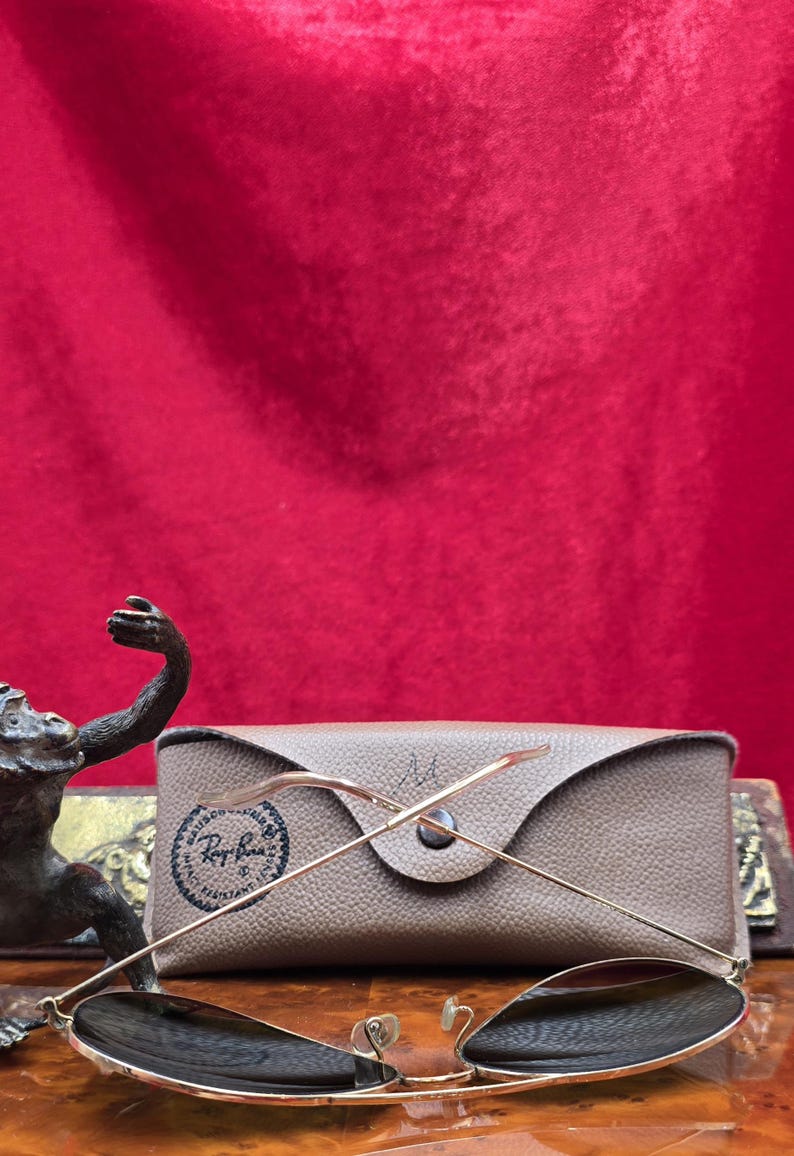 May include: Gold-framed sunglasses with dark lenses rest on a tan case. The case has a button closure and a logo. The sunglasses are positioned in front of the case, with the arms extended. The background is a solid red.