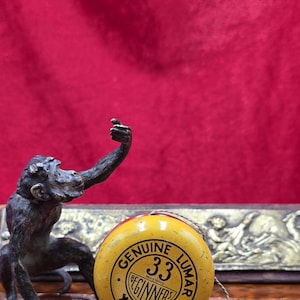 May include: A vintage yellow yo-yo with black text that reads "Genuine 33 Beginners Yo-Yo" next to a bronze monkey statue. The yo-yo has a string attached. The background is a deep red.