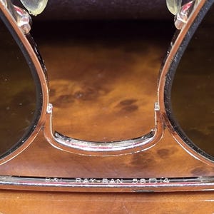 May include: Close-up of Ray-Ban sunglasses with a gold-colored frame and dark lenses. The text "RAY-BAN 58 14" is visible on the frame. The sunglasses are set against a brown, wood-grain background.