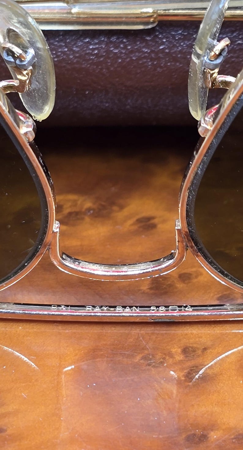 May include: Close-up of Ray-Ban sunglasses with a gold-colored frame and dark lenses. The text "RAY-BAN 58 14" is visible on the frame. The sunglasses are set against a brown, wood-grain background.