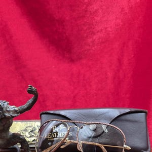 May include: Ray-Ban LEATHERS sunglasses with a brown leather frame and a black leather case. The sunglasses have a gold-coloured metal frame and clear lenses. The text "Ray-Ban LEATHERS" is visible on the lens.