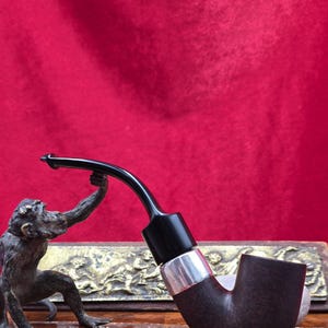 Pode incluir: Um cachimbo de tabaco castanho escuro com uma haste preta curva e uma faixa prateada. Uma estátua de bronze de um macaco está posicionada à esquerda, parecendo segurar o cachimbo. O fundo é vermelho liso.