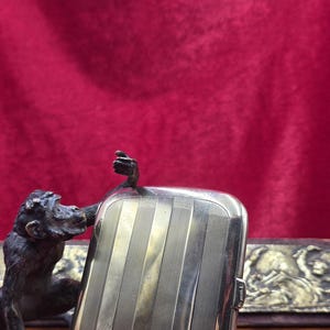 May include: A silver rectangular cigarette case with vertical lines, next to a bronze monkey statue. The case has a small clasp and is set against a red background. A second decorative item is visible in the background.