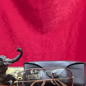 May include: A pair of Ray-Ban Leathers sunglasses with a brown leather frame and gold-tone metal detailing, alongside a black leather case. The sunglasses feature a classic aviator design. The text "Ray-Ban Leathers BAUSCH & LOMB" is visible.
