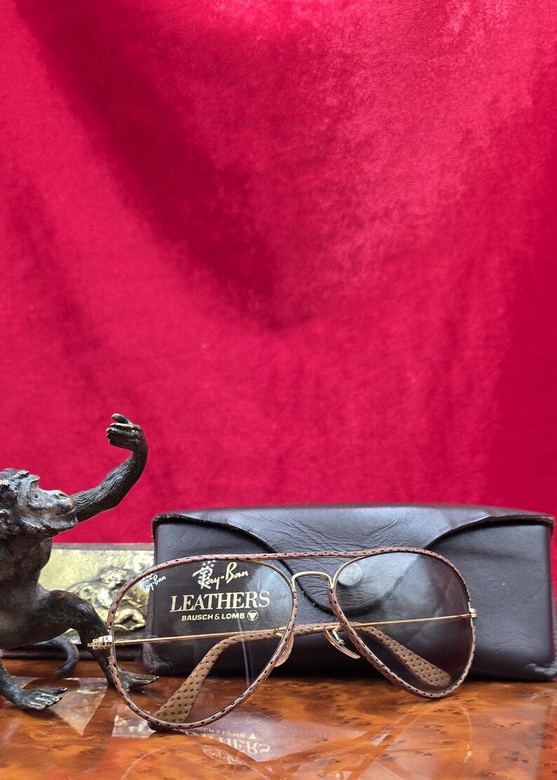 May include: A pair of Ray-Ban Leathers sunglasses with a brown leather frame and gold-tone metal detailing, alongside a black leather case. The sunglasses feature a classic aviator design. The text "Ray-Ban Leathers BAUSCH & LOMB" is visible.