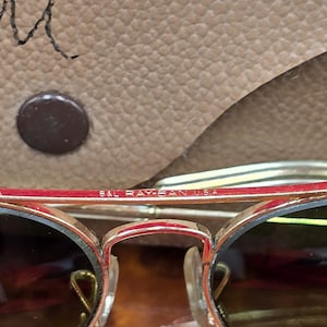 May include: Close-up of gold-rimmed sunglasses with dark lenses. The frame has a red accent and the text "B&L RAY-BAN U.S.A." is visible. The sunglasses are set against a tan leather case.