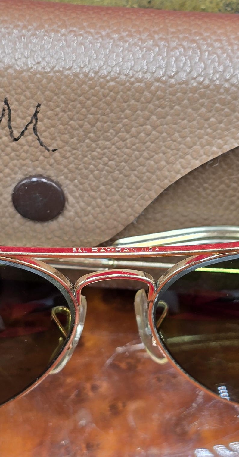 May include: Close-up of gold-rimmed sunglasses with dark lenses. The frame has a red accent and the text "B&L RAY-BAN U.S.A." is visible. The sunglasses are set against a tan leather case.
