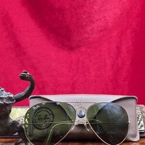May include: Gold-framed aviator sunglasses with green lenses, resting on a tan case. The sunglasses have a classic design with a double bridge and clear nose pads. The case is rectangular and has a smooth texture. The background is a solid red.