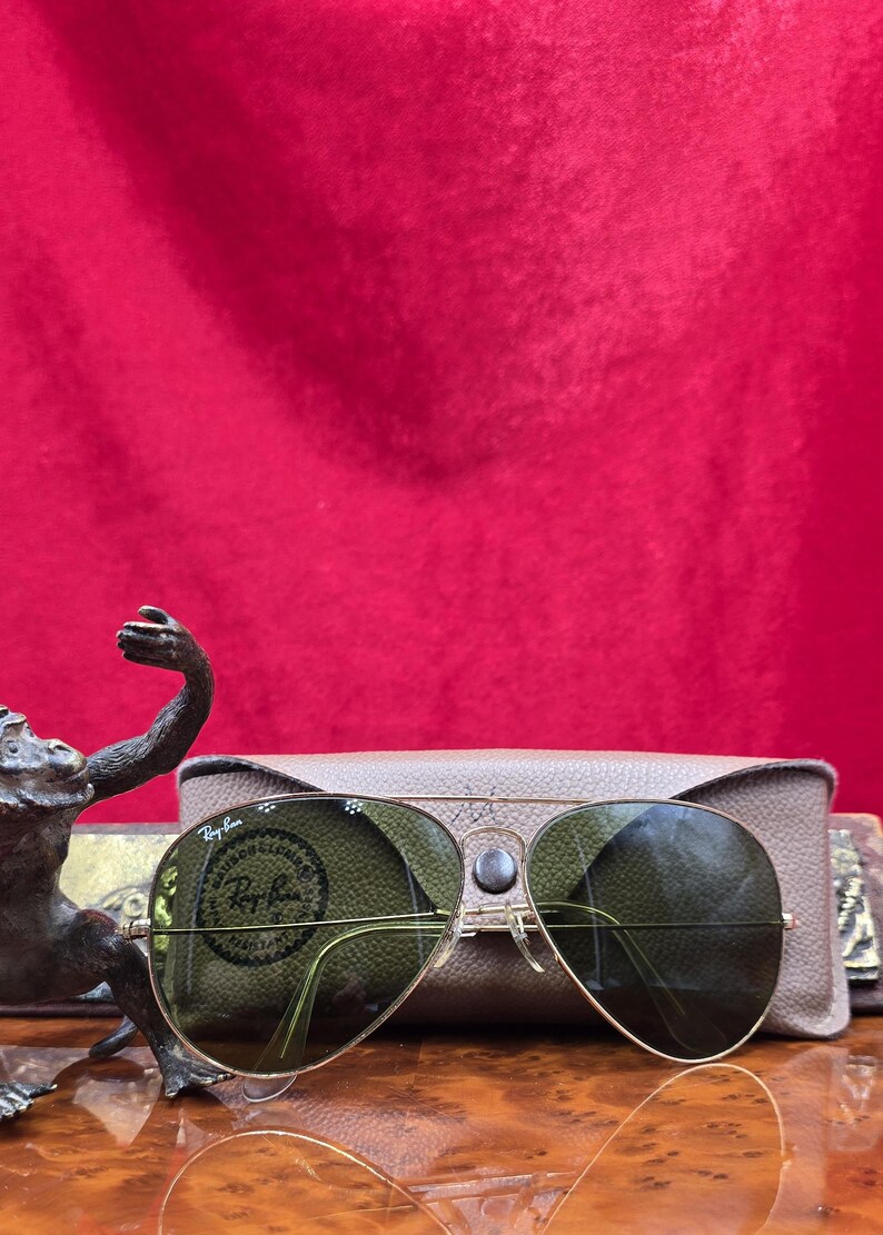 May include: Gold-framed aviator sunglasses with green lenses, resting on a tan case. The sunglasses have a classic design with a double bridge and clear nose pads. The case is rectangular and has a smooth texture. The background is a solid red.