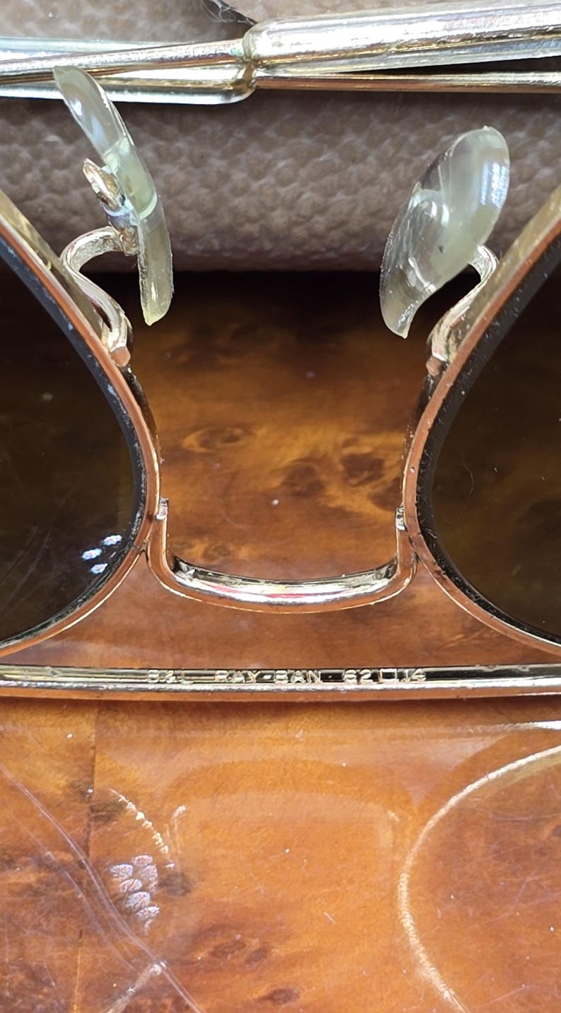 May include: Close-up of gold-framed sunglasses with dark lenses. The Ray-Ban logo is visible on the frame. The sunglasses have clear nose pads and a classic aviator design.