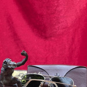 May include: A bronze monkey statue with an outstretched arm sits next to a pair of gold-rimmed sunglasses with dark lenses. A brown leather glasses case is behind the sunglasses. The background is a deep red.