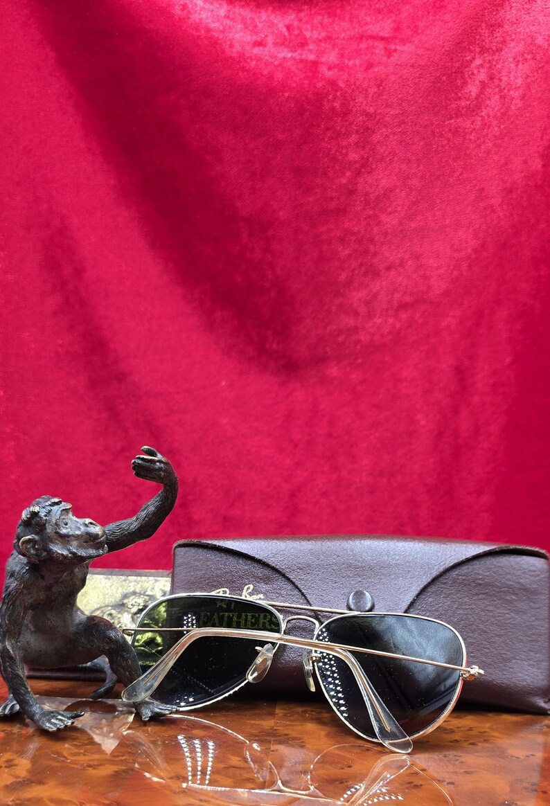 May include: A bronze monkey statue with an outstretched arm sits next to a pair of gold-rimmed sunglasses with dark lenses. A brown leather glasses case is behind the sunglasses. The background is a deep red.