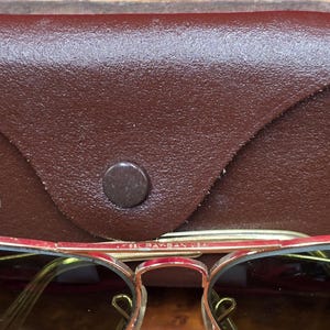 May include: Close-up of vintage sunglasses with a brown leather case. The sunglasses have a gold-colored frame with red accents and brown lenses. The case is a dark brown color with a button closure. The text "B&L RAY-BAN USA" is visible on the frame.