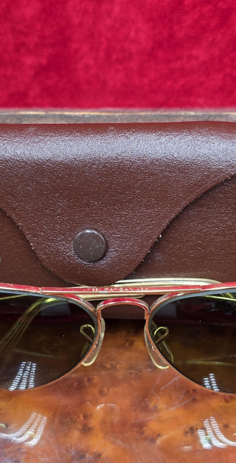 May include: Close-up of vintage sunglasses with a brown leather case. The sunglasses have a gold-colored frame with red accents and brown lenses. The case is a dark brown color with a button closure. The text "B&L RAY-BAN USA" is visible on the frame.