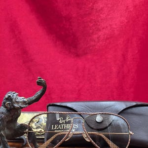 May include: Ray-Ban aviator sunglasses with brown leather detailing and a matching case. The sunglasses have a gold frame and brown lenses. The case is black leather. A bronze monkey statue is in the background.