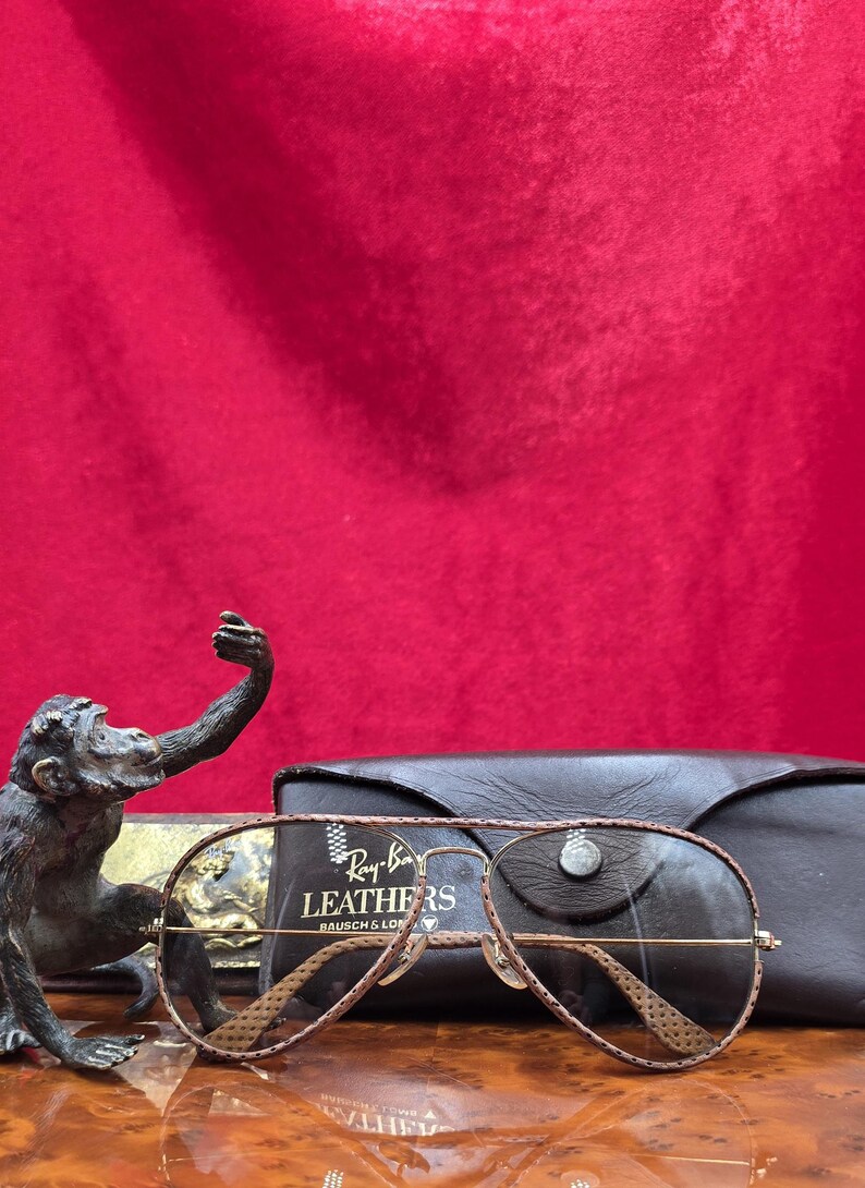 May include: Ray-Ban aviator sunglasses with brown leather detailing and a matching case. The sunglasses have a gold frame and brown lenses. The case is black leather. A bronze monkey statue is in the background.