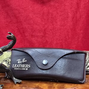 May include: A dark brown leather glasses case with a snap closure. The case features the Ray-Ban Leathers logo in white. It rests on a wooden surface against a red backdrop.