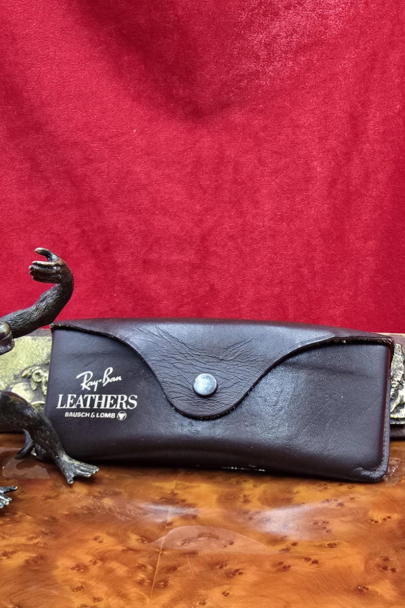 May include: A dark brown leather glasses case with a snap closure. The case features the Ray-Ban Leathers logo in white. It rests on a wooden surface against a red backdrop.
