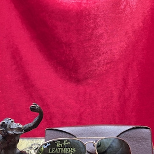 May include: Gold-framed Ray-Ban Leathers sunglasses with green lenses, resting on a brown leather case. A bronze monkey statue is in the foreground. The background is a deep red velvet.