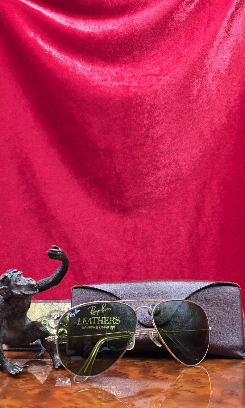 May include: Gold-framed Ray-Ban Leathers sunglasses with green lenses, resting on a brown leather case. A bronze monkey statue is in the foreground. The background is a deep red velvet.