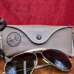 May include: Gold-framed aviator sunglasses with dark lenses rest on a wooden surface, next to a textured, tan leather case. The case features a black button closure and a circular logo. The sunglasses are a classic style, suitable for sun protection.