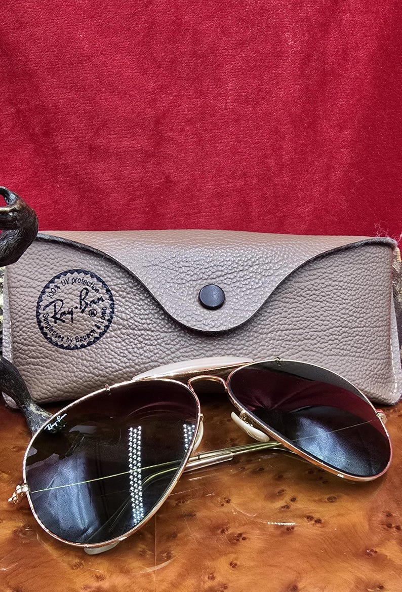 May include: Gold-framed aviator sunglasses with dark lenses rest on a wooden surface, next to a textured, tan leather case. The case features a black button closure and a circular logo. The sunglasses are a classic style, suitable for sun protection.