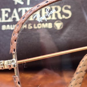 May include: Close-up of Ray-Ban sunglasses with brown leather detailing. The lenses are a light brown tint, and the frame is gold. The Ray-Ban logo is visible on the lens and the case.