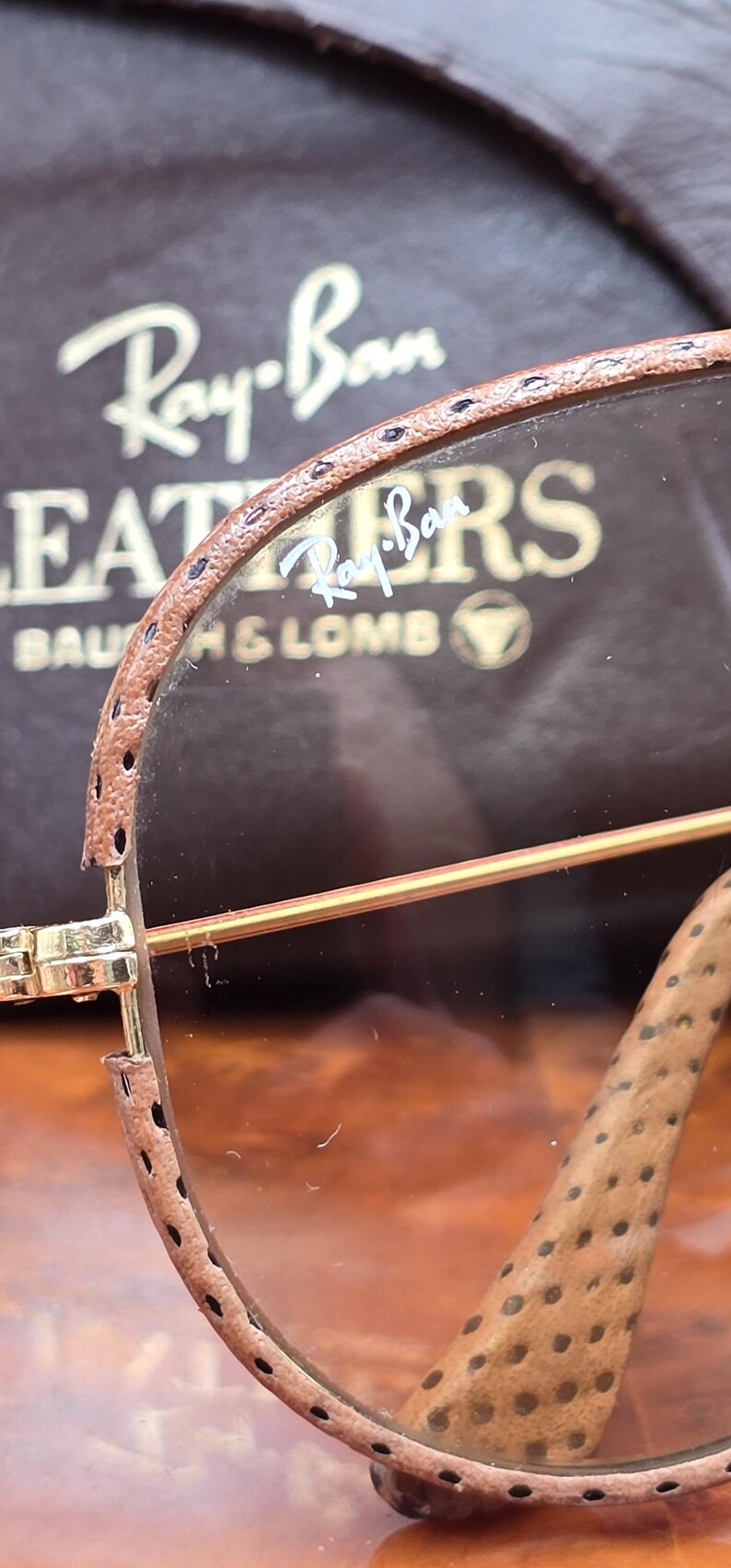 May include: Close-up of Ray-Ban sunglasses with brown leather detailing. The lenses are a light brown tint, and the frame is gold. The Ray-Ban logo is visible on the lens and the case.