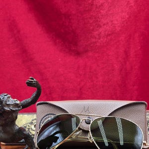 May include: Gold-rimmed aviator sunglasses with green lenses rest on a wooden surface, next to a tan leather case. A bronze monkey statue is in the foreground. The background is a red velvet cloth.