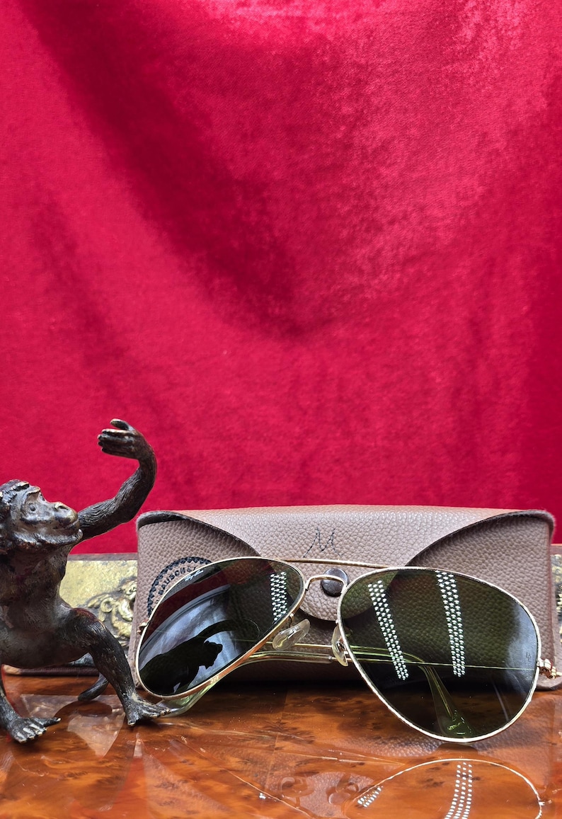 May include: Gold-rimmed aviator sunglasses with green lenses rest on a wooden surface, next to a tan leather case. A bronze monkey statue is in the foreground. The background is a red velvet cloth.