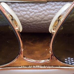 May include: Close-up of gold-rimmed sunglasses with dark lenses. The sunglasses have a classic design with a bridge and arms. The lenses are a dark shade, and the frames are gold. The sunglasses are resting on a light brown surface.