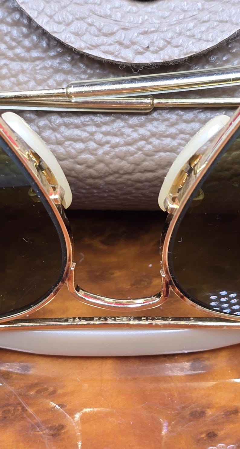 May include: Close-up of gold-rimmed sunglasses with dark lenses. The sunglasses have a classic design with a bridge and arms. The lenses are a dark shade, and the frames are gold. The sunglasses are resting on a light brown surface.