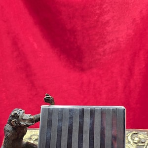 May include: A silver cigarette case with vertical striped detailing. A bronze monkey statue is positioned to the left of the case, with its arm raised. The background is a solid red.