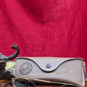 May include: Gold-framed sunglasses with dark lenses rest in front of a brown leather case. The case has a button closure and a logo. The sunglasses are on a wooden surface against a red background.
