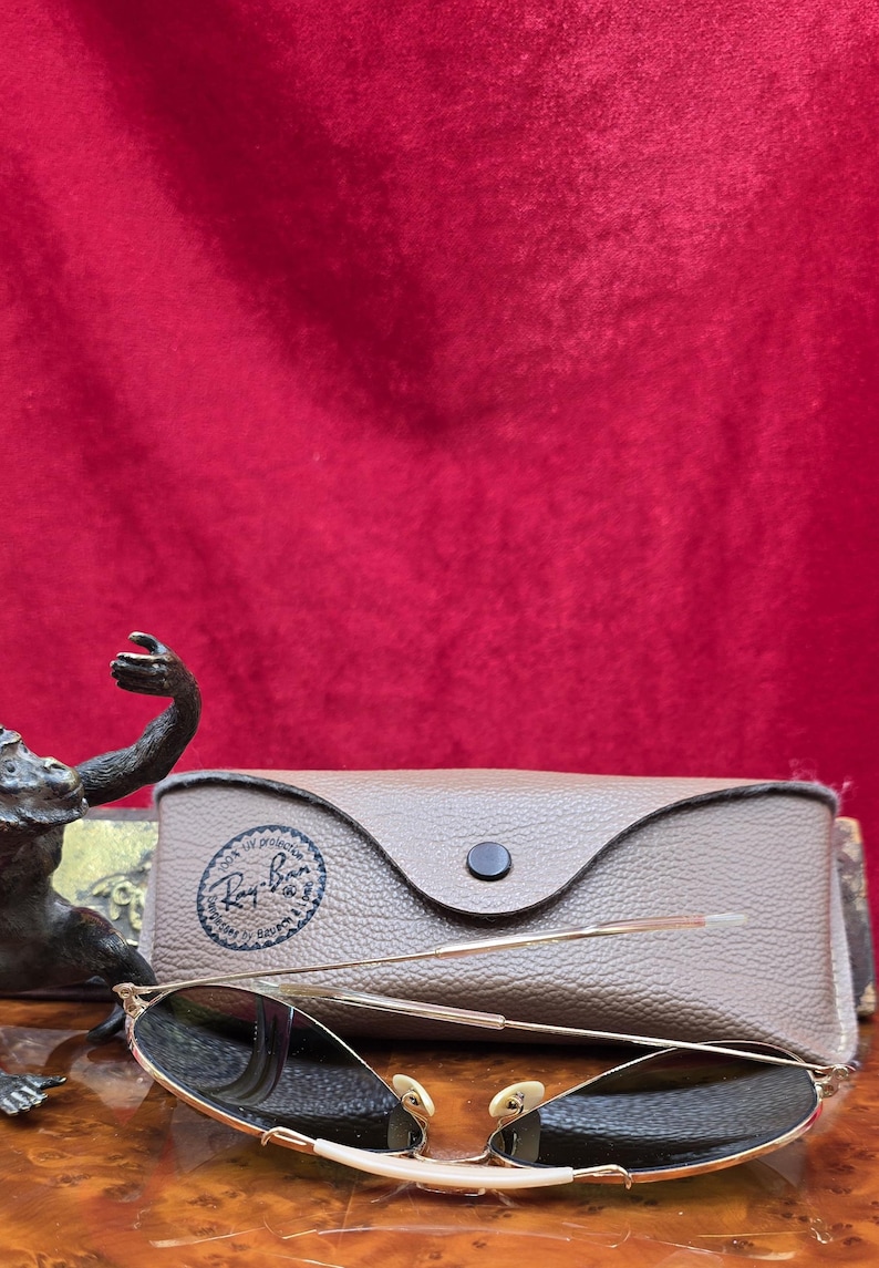 May include: Gold-framed sunglasses with dark lenses rest in front of a brown leather case. The case has a button closure and a logo. The sunglasses are on a wooden surface against a red background.