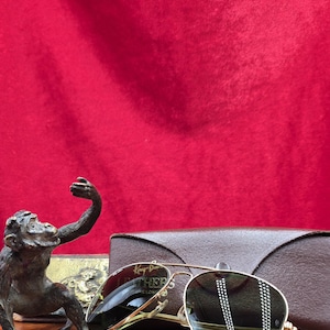 May include: Gold-framed sunglasses with dark lenses, resting on a wooden surface. The sunglasses are next to a brown leather case and a bronze monkey statue. The background is a solid red fabric.