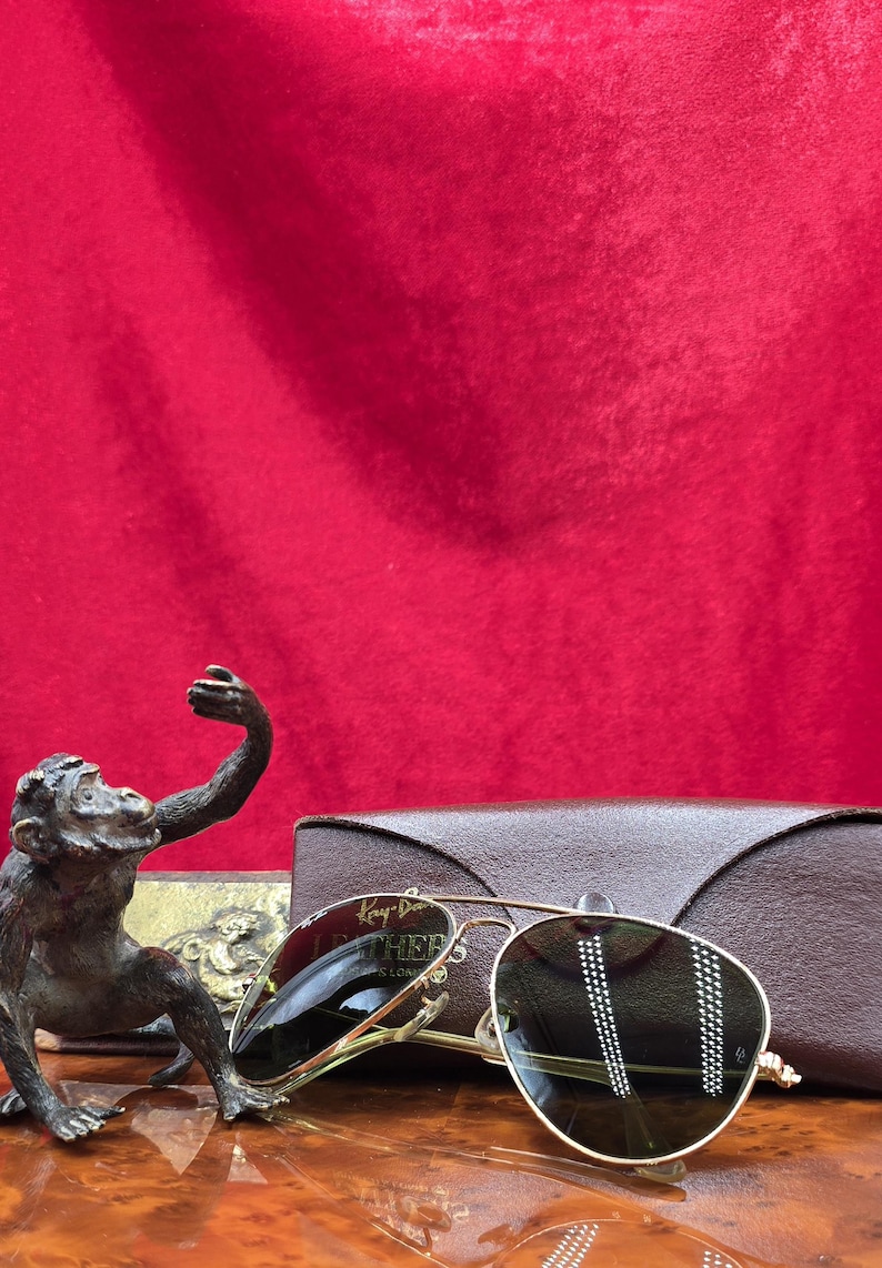 May include: Gold-framed sunglasses with dark lenses, resting on a wooden surface. The sunglasses are next to a brown leather case and a bronze monkey statue. The background is a solid red fabric.