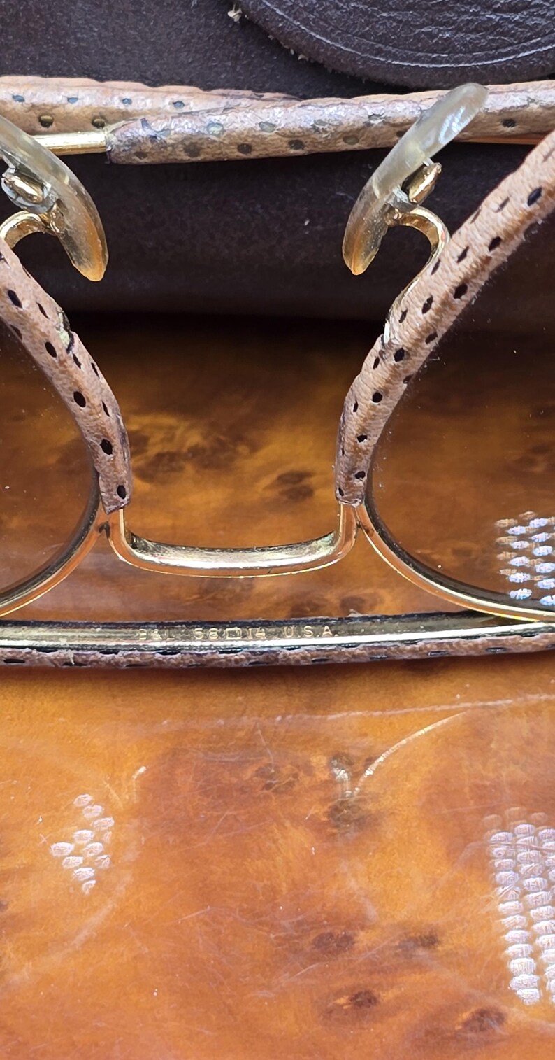 May include: Close-up of vintage sunglasses with gold frames and brown lenses. The temples are covered in a spotted, light brown material. The text "BAL 58 14 USA" is visible on the frame.