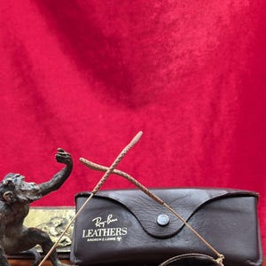 May include: Ray-Ban Leathers sunglasses with a brown leather case. The sunglasses have a gold frame and brown lenses. The case is dark brown with the Ray-Ban Leathers logo. A bronze monkey statue is in the background.