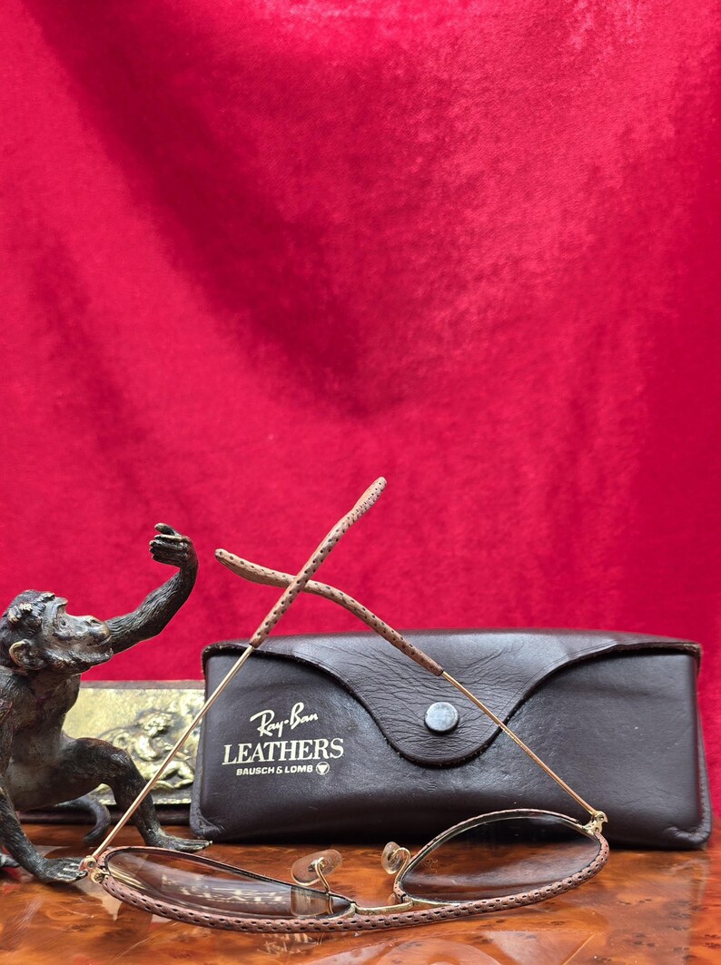 May include: Ray-Ban Leathers sunglasses with a brown leather case. The sunglasses have a gold frame and brown lenses. The case is dark brown with the Ray-Ban Leathers logo. A bronze monkey statue is in the background.