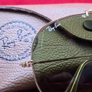 May include: Close-up of Ray-Ban sunglasses with a gold frame and green lenses. The Ray-Ban logo is visible on the lens. A brown leather case is in the background. The sunglasses are on a wooden surface.