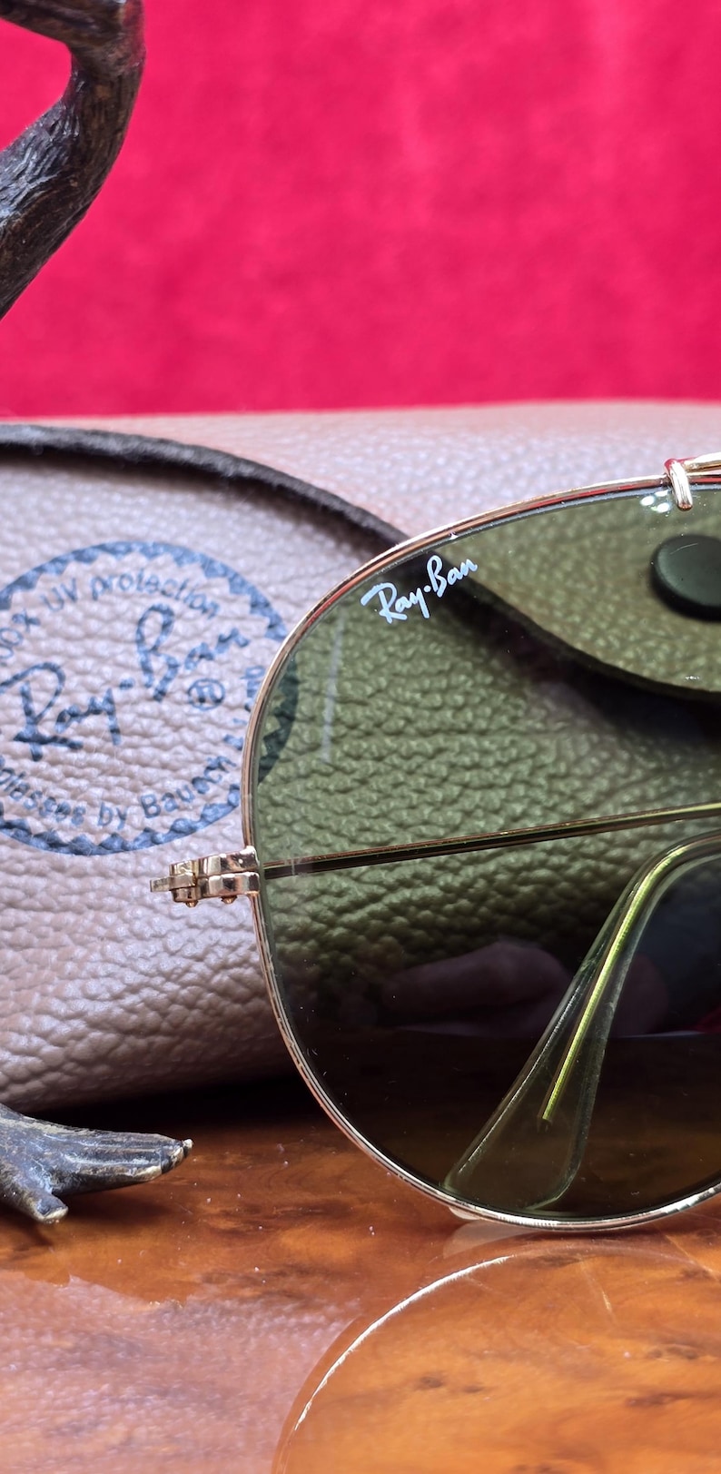 May include: Close-up of Ray-Ban sunglasses with a gold frame and green lenses. The Ray-Ban logo is visible on the lens. A brown leather case is in the background. The sunglasses are on a wooden surface.