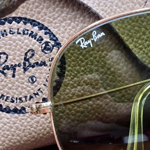 May include: Close-up of Ray-Ban sunglasses with a gold frame and green lenses. The Ray-Ban logo is visible on the lens. The sunglasses are resting on a tan leather case. The case has the Ray-Ban logo and text.