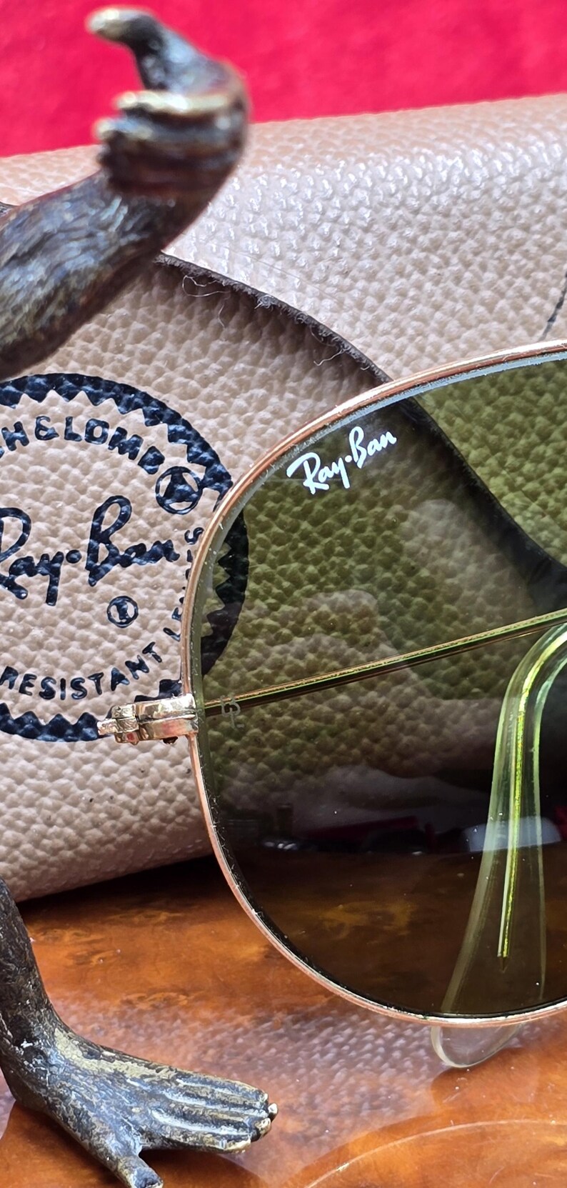 May include: Close-up of Ray-Ban sunglasses with a gold frame and green lenses. The Ray-Ban logo is visible on the lens. The sunglasses are resting on a tan leather case. The case has the Ray-Ban logo and text.