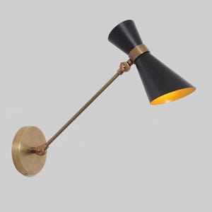 Könnte beinhalten: Schwarz-goldene Wandleuchte mit konischem Schirm und messingfarbenem Arm und Sockel. Das Innere des Schirms ist goldfarben, und die Leuchte ist verstellbar.