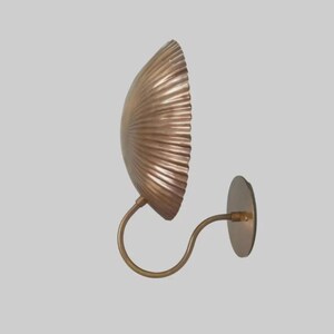 May include: A wall-mounted sconce with a fan-shaped shade and a curved arm. The shade has a textured, ridged surface and a warm, metallic finish. The sconce is attached to a round base.