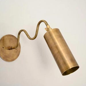 May include: A brass wall sconce with a cylindrical light fixture and a curved arm. The sconce has a round base and is mounted on a white wall. The light fixture is angled downward.