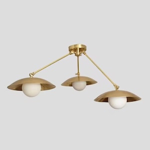 May include: A brass-toned ceiling light fixture with three adjustable arms. Each arm supports a shallow, circular shade with a white globe light. The fixture has a modern, minimalist design.