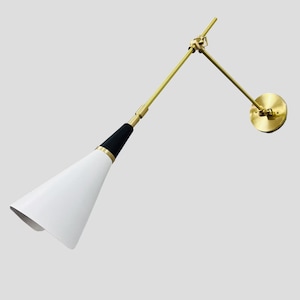Può includere: Una lampada da parete moderna con un paralume conico bianco e un dettaglio nero. La lampada presenta un braccio regolabile color ottone e un supporto a parete circolare. Il design è elegante e minimalista, adatto a vari stili di interni.