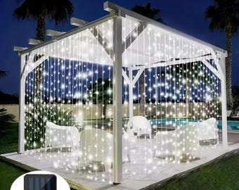 May include: White solar powered LED curtain lights strung across a white pergola with a solar panel on the ground. The lights are on and illuminating the pergola.